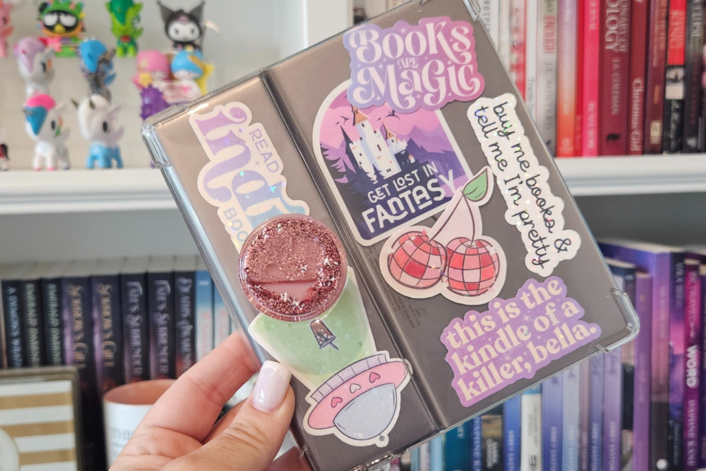 A colorful bookshelf filled with books is the backdrop as Sarra holds up a silver Kindle Oasis with a clear cover and many colorful stickers.